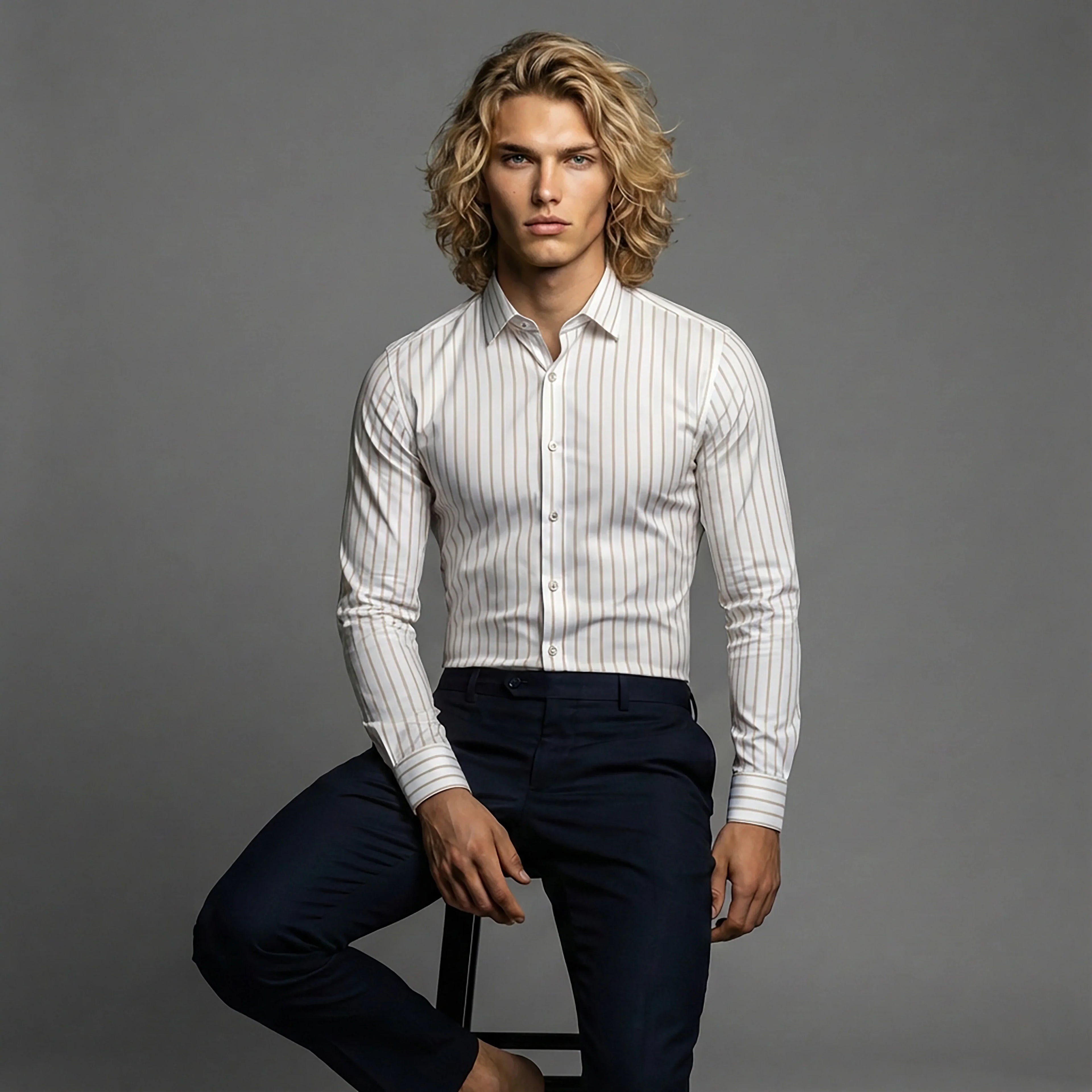 Man in DGMAH long sleeve white striped shirt and navy pants sitting on stool, studio background